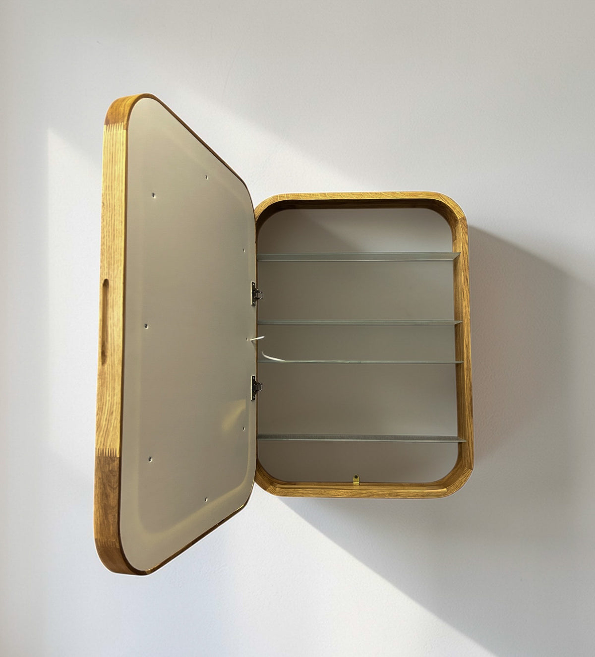 A rectangular wall-mounted wooden storage cabinet with an open hinged door, featuring three glass shelves inside, shown against a plain white wall.