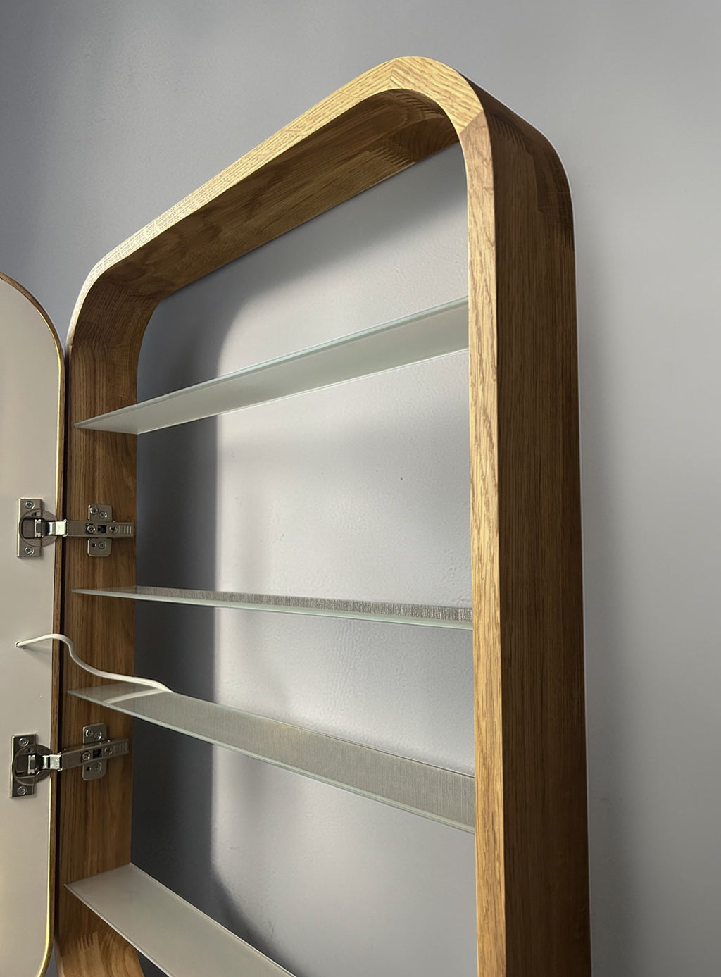 A close-up of a wall-mounted wooden cabinet or shelving unit with a curved top, three glass shelves, and visible metal hinges against a gray wall.
