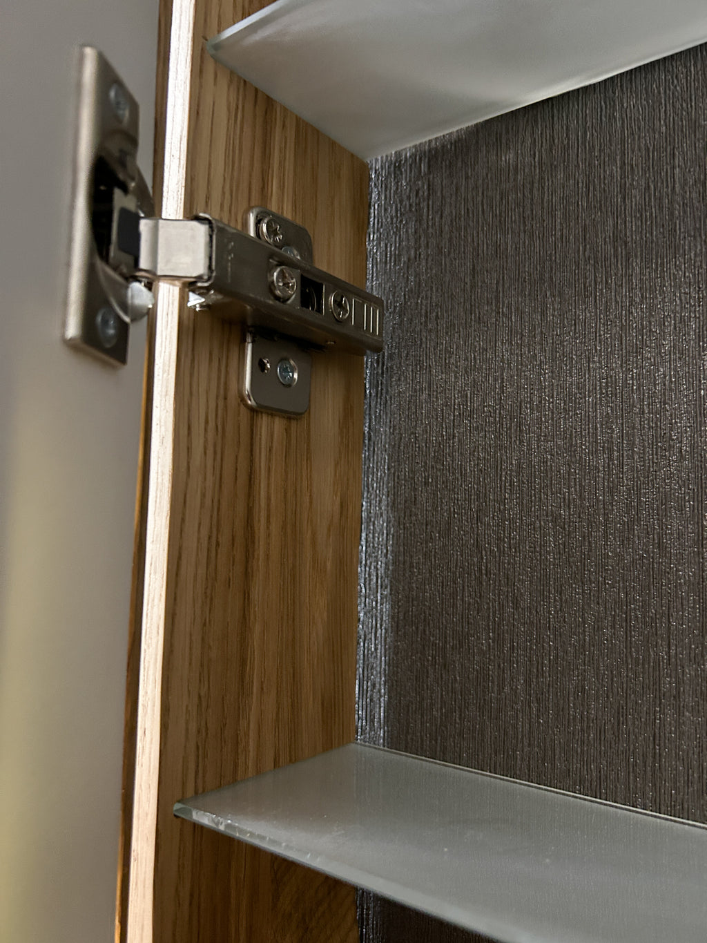 Close-up of a cabinet door hinge mounted on a wood-grain cabinet door, with a textured dark gray interior wall and a shelf visible.