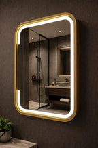 A rectangular wall mirror with rounded corners, a warm gold frame, and integrated LED lighting, mounted on a dark textured bathroom wall. The mirror reflects a modern bathroom interior with a glass shower enclosure, wooden vanity, and vessel sink.
