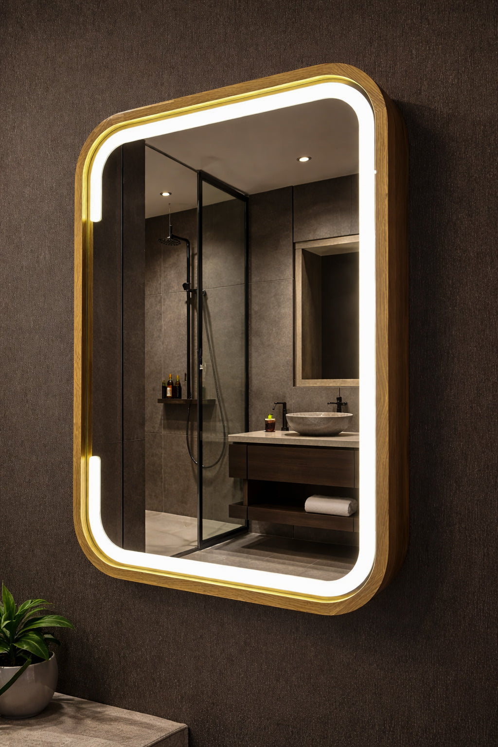 A rectangular wall mirror with rounded corners, a warm gold frame, and integrated LED lighting, mounted on a dark textured bathroom wall. The mirror reflects a modern bathroom interior with a glass shower enclosure, wooden vanity, and vessel sink.