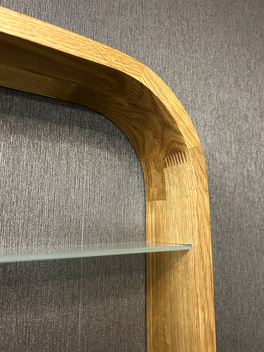 Close-up of a wooden shelf frame with a curved top and glass shelf, set against a textured gray wall.