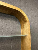 Close-up of a wooden shelf frame with a curved top and glass shelf, set against a textured gray wall.