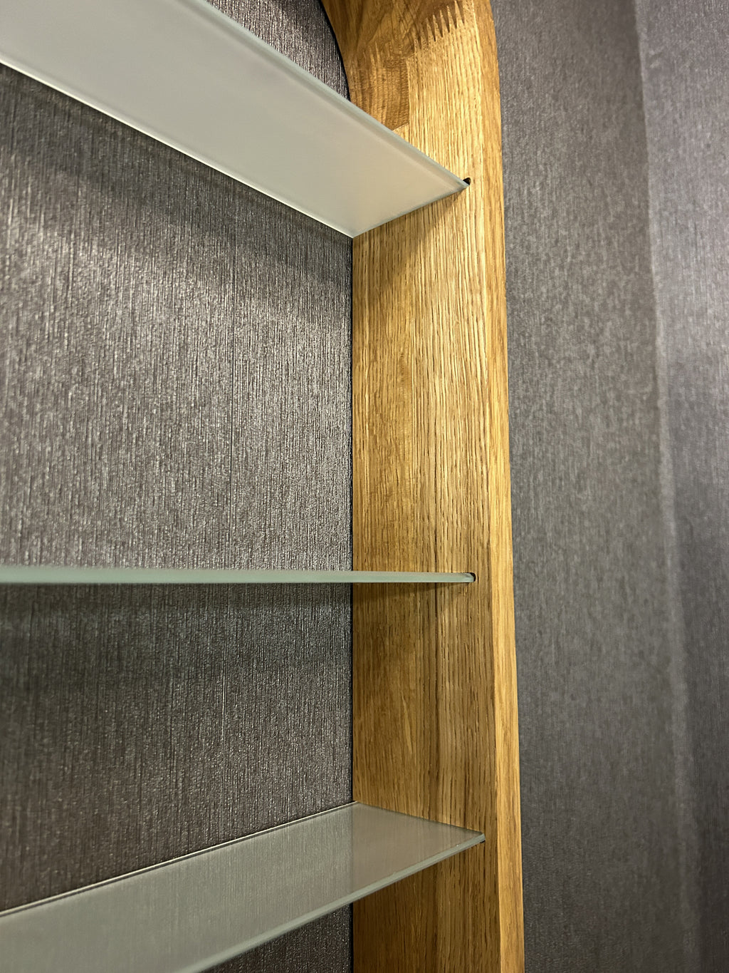 Close-up of a built-in shelving unit with three glass shelves and a light natural wood frame, set against a gray textured wall.