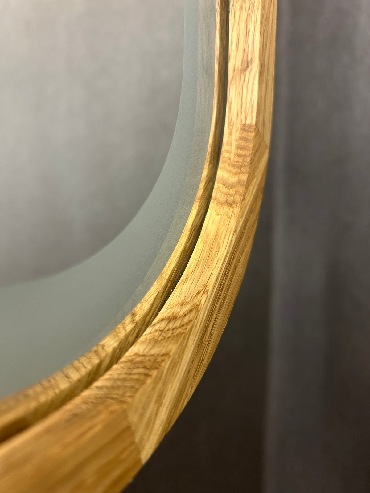 Close-up detail of a round mirror with a curved light oak wood frame, showing the natural wood grain and the edge of the frosted glass.