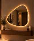 Asymmetrical pebble-shaped backlit LED mirror, frameless, above a floating wood console