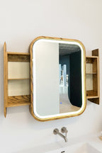 A wall-mounted medicine cabinet with a light oak wood frame, built-in side shelves, and an illuminated LED light border around the mirror, shown mounted on a white wall above a sink.