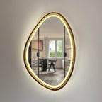 An organic-shaped wall mirror with a warm glowing LED light border and a metallic gold frame, mounted on a light beige wall. The reflection shows a modern home office interior.