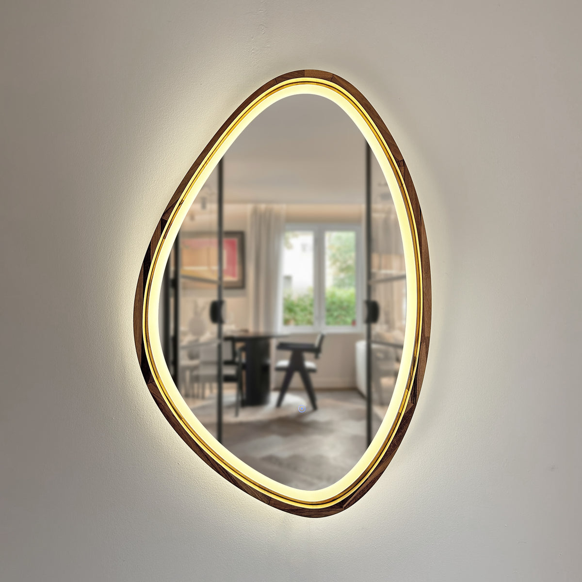An organic-shaped wall mirror with a warm glowing LED light border and a metallic gold frame, mounted on a light beige wall. The reflection shows a modern home office interior.
