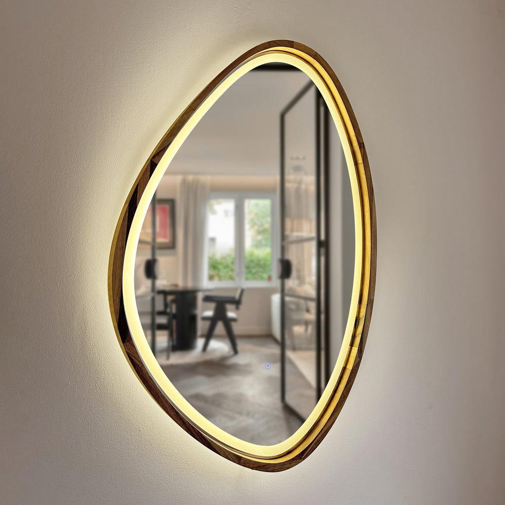 An organic-shaped wall mirror with a brushed gold frame and integrated warm white LED lighting, mounted on a textured off-white wall. The mirror reflects a modern room interior with a desk and chair.
