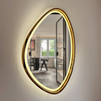 An organic-shaped wall mirror with a brushed gold frame and integrated warm white LED lighting, mounted on a textured off-white wall. The mirror reflects a modern room interior with a desk and chair.