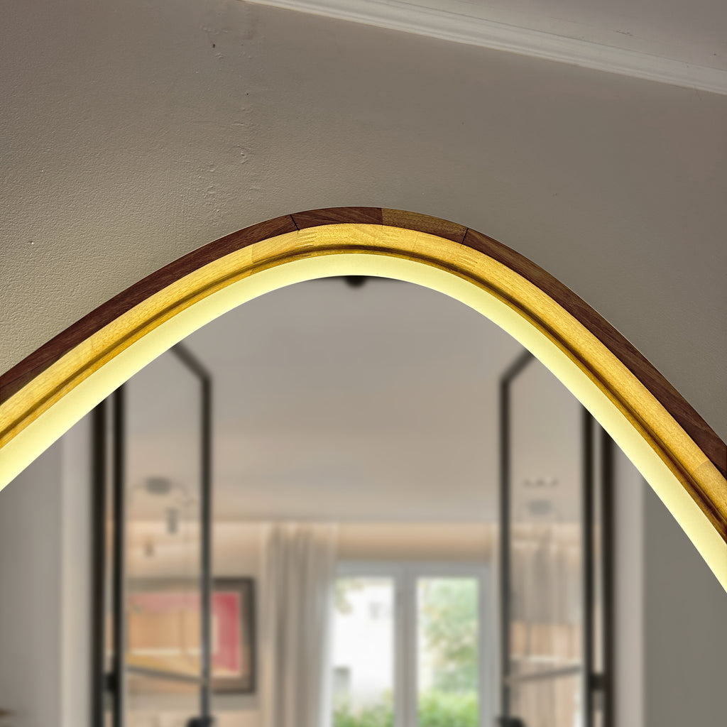 An arched wall mirror with a warm glowing LED light strip integrated into a layered frame of dark brown wood and light wood grain, shown in a home interior setting.