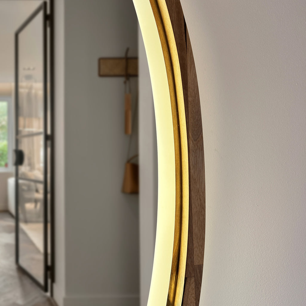 A close-up of a modern round wall mirror with a warm white LED light ring and a dark wood frame, shown in a minimalist hallway setting.