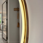 A close-up of a modern round wall mirror with a warm white LED light ring and a dark wood frame, shown in a minimalist hallway setting.