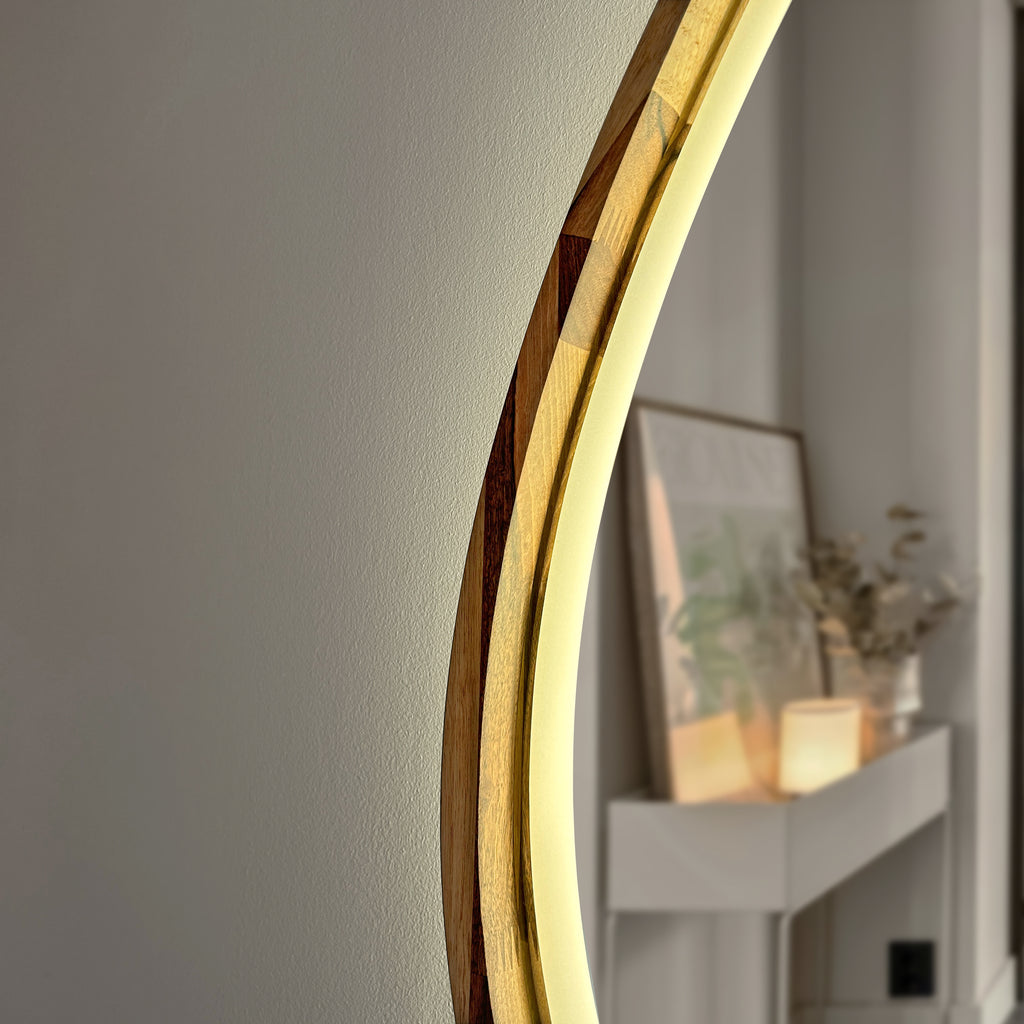 A close-up of a modern curved wall mirror with a warm wood and gold-toned frame, reflecting a softly lit interior shelf with framed art and candles.