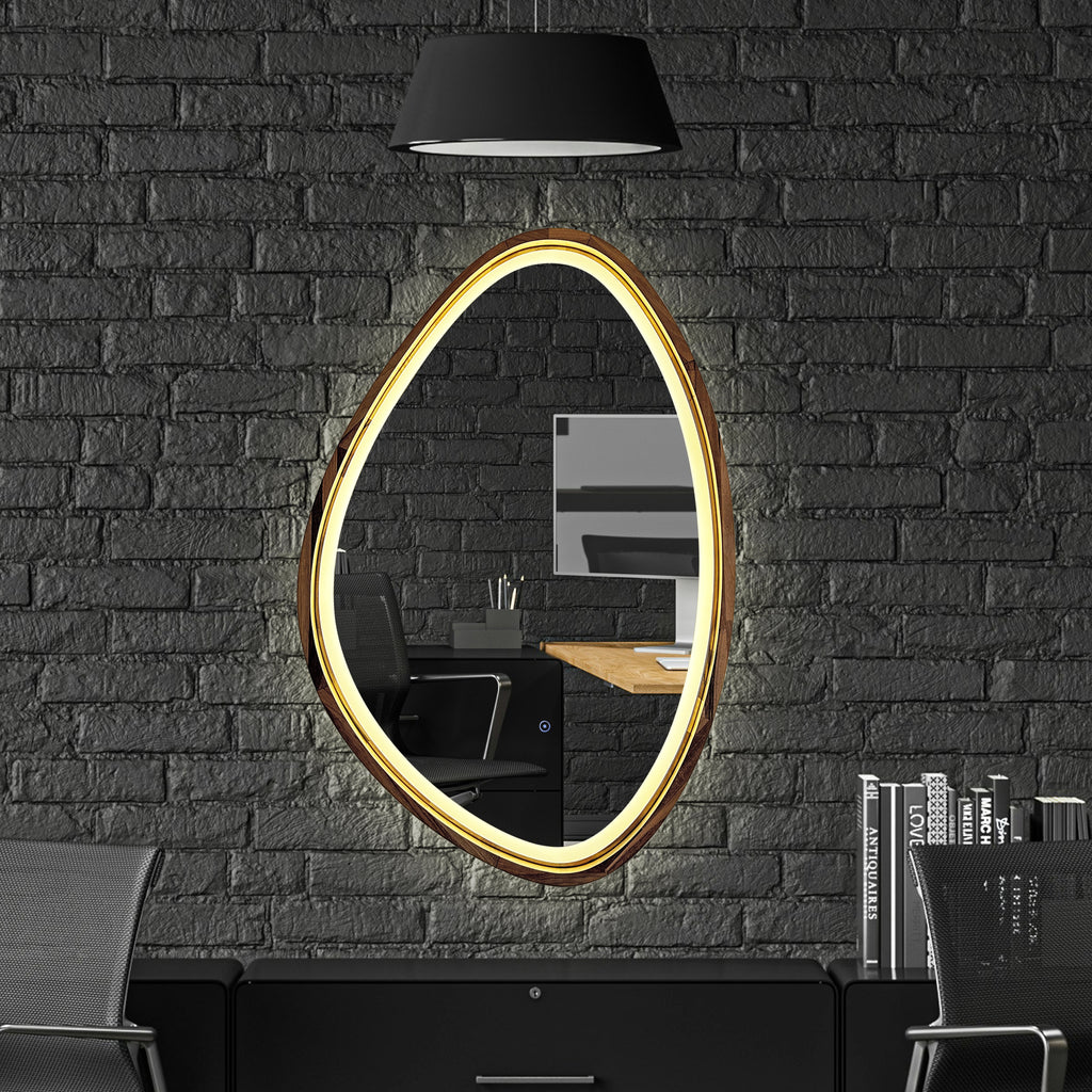 An oval wall mirror with a glowing warm gold LED frame, mounted on a dark gray brick wall above a black desk setup with a chair and a black pendant lamp.
