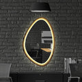 An oval wall mirror with a glowing warm gold LED frame, mounted on a dark gray brick wall above a black desk setup with a chair and a black pendant lamp.