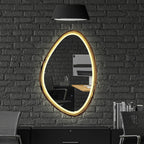 An oval wall mirror with a glowing warm gold LED frame, mounted on a dark gray brick wall above a black desk setup with a chair and a black pendant lamp.