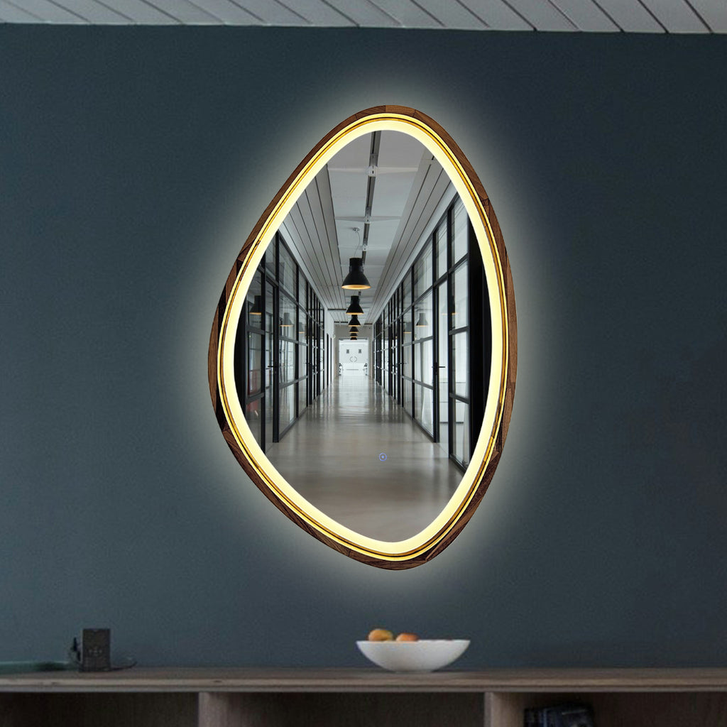 An organic-shaped LED wall mirror mounted on a dark slate blue-gray wall above a wooden console table.
