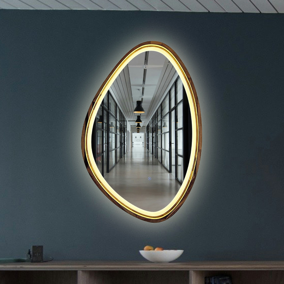 An organic-shaped LED wall mirror mounted on a dark slate blue-gray wall above a wooden console table.
