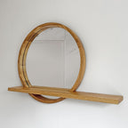 A round wall mirror with a thick natural oak wood frame and a matching floating shelf, mounted on a plain white wall.