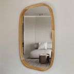 An organic-shaped wall mirror with a natural light oak wood frame, mounted on a neutral beige wall. The mirror reflects a modern living room interior featuring a light gray sofa, a metallic side table, and a ribbed wall background.