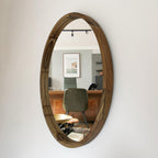 An oval wooden wall mirror with a rustic, handcrafted dark brown frame, mounted on a plain white wall. The mirror reflects a modern office interior with a desk, chair, and framed art.