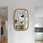 A vertical oval wall mirror with a natural bamboo wood frame, mounted on a white wall in a modern hallway. The mirror reflects a dining table with a vase of yellow flowers, a black pendant light, and a kitchen in the background.