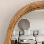 A close-up of a large arched wooden mirror frame in a modern living room setting, reflecting a light gray sofa and decorative wall art.