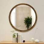 A round wall mirror with a rustic dark brown wooden frame hangs on a light beige wall above a wooden console table styled with books, small potted plants, and ceramic vases.