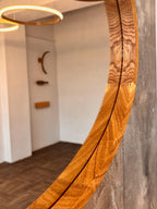 A close-up of a large, organic-shaped wooden mirror with a rich grain pattern, mounted on a textured gray wall in a modern interior setting.