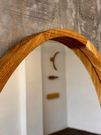 A close-up of a curved wooden mirror frame with a warm honey-brown finish, set against a textured gray wall and reflecting a softly blurred interior room.