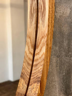 Close-up of a curved wooden chair or stool leg with prominent natural wood grain, positioned against a textured gray wall and a blurred indoor background.