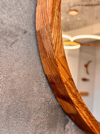 Close-up of a curved wooden mirror frame with a rich, warm honey-brown wood grain pattern, set against a textured gray wall with a softly blurred interior reflection.