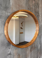 Round wooden wall mirror with a thick natural oak frame, mounted on a textured gray concrete wall. The mirror reflects a hallway with white walls and a gold ring light.