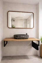 A modern wall-mounted bathroom vanity with a light wood shelf, black metal brackets, a dark stone vessel sink, and a large rectangular mirror with a warm wood frame on a light beige tiled wall.